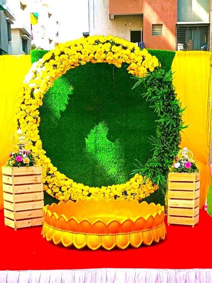 A simple Haldi decoration for an outdoor space. The setup features a circular frame of marigolds against a faux grass wall, with a lotus-shaped seat.