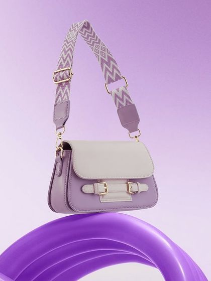 A trendy lavender handbag with a patterned strap, balanced on a swirling purple prop. The monochromatic color scheme is modern and playful.