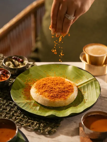 A hand sprinkling podi masala over a soft idli. This action shot highlights the fresh spices and adds a dynamic element to the photo.
