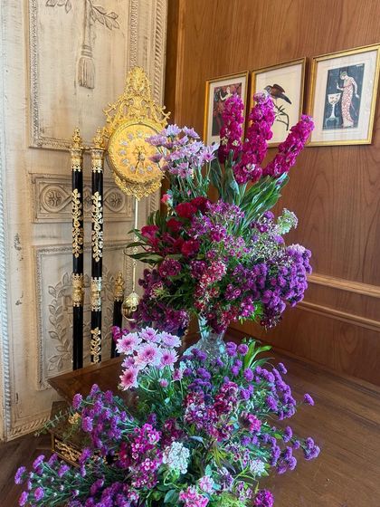 An arrangement of pink and purple florals stands beside an antique clock, blending modern vibrancy with classic elegance.