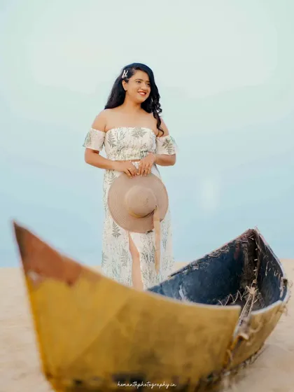 A beautiful solo portrait of the bride-to-be standing on the beach next to a rustic boat, holding a sun hat.