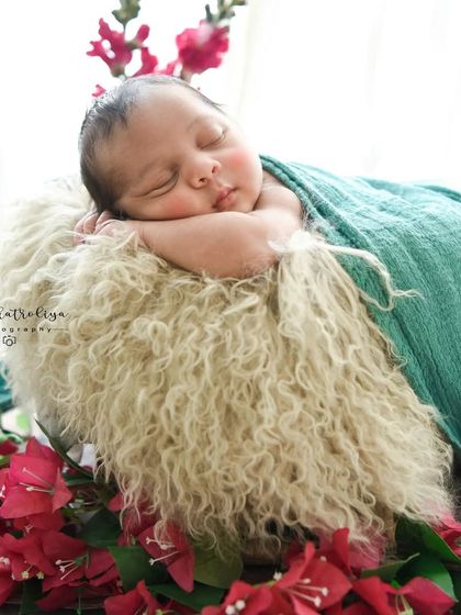 A different pose to capture their delicate features. Here, the baby rests on their tummy on a fluffy rug, draped in a teal wrap and framed by bright pink bougainvillea.