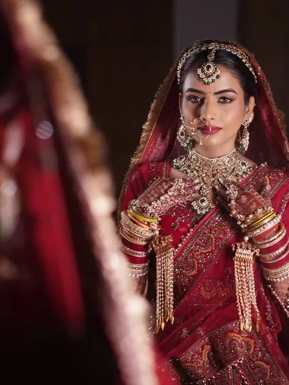 Adjusting the final details. The rich red lehenga is complemented by classic makeup with defined eyes and a warm, glowing complexion, ensuring the bride looks and feels her absolute best.