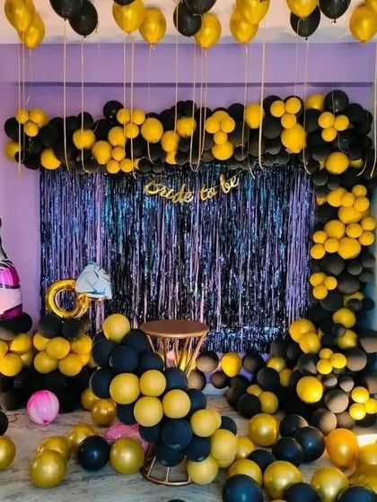 A fun and glamorous 'Bride to be' party in black and yellow. The decor features a fringe curtain backdrop, balloon columns, and fun accents like a champagne bottle and ring balloon.