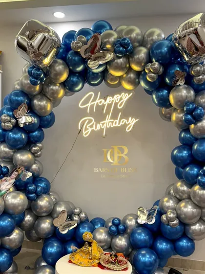 An enchanting Janmashtami or birthday balloon backdrop. This full ring of metallic blue and silver balloons is accented with butterfly details and a 'Happy Birthday' neon sign, with a Krishna idol at the center.