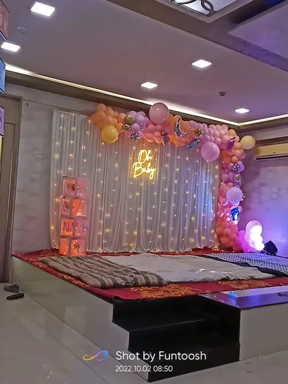 A stage setup for a baby shower, or 'Dohale Jevan', in a banquet hall. It features a balloon arch and a fairy light backdrop with an 'Oh Baby' neon sign.
