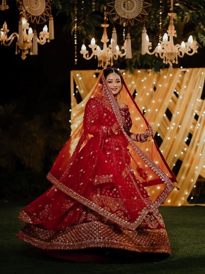 A beautiful shot of the bride twirling in her red wedding lehenga. The outdoor setting and the warm lights of the chandeliers create a magical, fairytale-like atmosphere.