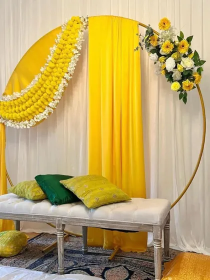A modern and elegant Haldi backdrop. This setup features a golden circular arch draped with bright yellow fabric and adorned with garlands of marigolds and white flowers.