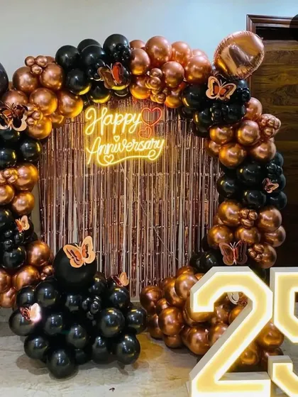 Here is another take on a 25th-anniversary celebration, using a modern color scheme. The setup includes a full balloon ring of black and rose gold chrome balloons, a rose gold fringe curtain, a "Happy Anniversary" neon sign, and illuminated "25" numbers.