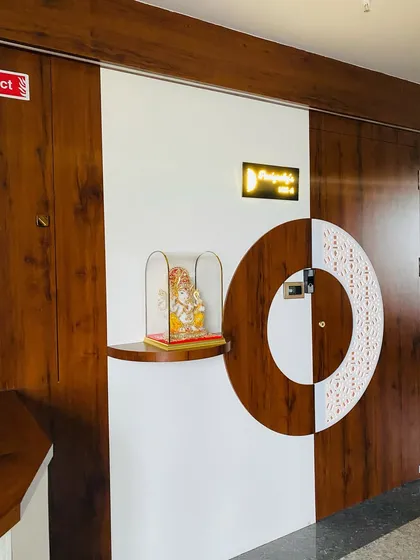 A unique entrance door design where two contrasting finishes, wood and white, meet in a circular pattern. A small shelf holds a Ganesha idol, blending modern design with tradition.