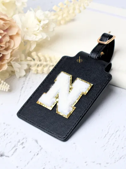 A single personalized luggage tag in black with a white and gold chenille "N" patch. These tags make it easy to spot your bag on the carousel.