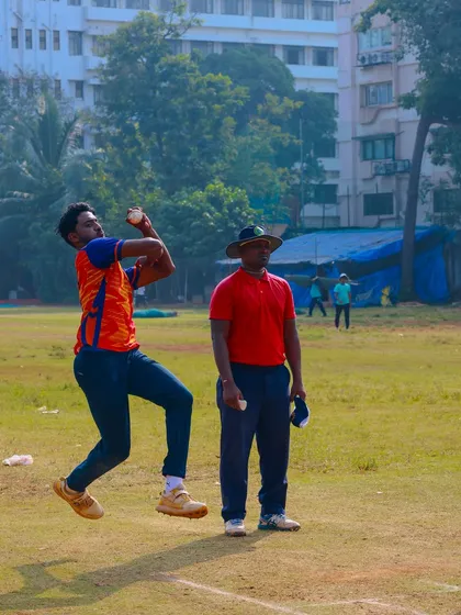 A fast bowler mid-air during his delivery stride in a practice match, watched by an umpire.