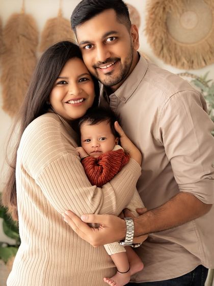 A happy family portrait with warm, earthy tones. The parents are holding their baby and smiling brightly for the camera.