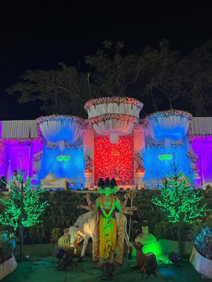 A spectacular wedding stage featuring a central red floral panel flanked by blue-lit sections and grand floral chandeliers. In the foreground, a statue of Lord Khandoba adds a traditional and divine element, perfect for a Koli-themed wedding.
