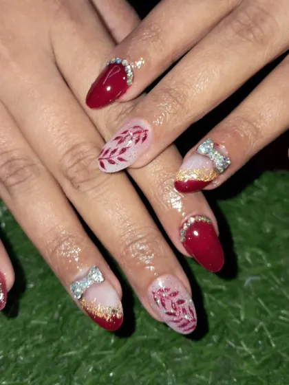 A close-up of a beautiful bridal nail set. We combined solid red nails, red glitter nails, and accent nails with delicate hand-painted leaves and 3D stone bows.