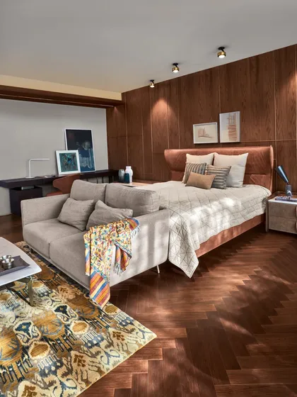 This bedroom in the Baid Residence is defined by its herringbone wood flooring and a custom bed with an integrated sofa. Natural light floods the space, highlighting the textures of the rug and linens.