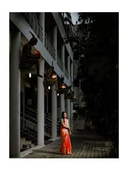 An artistic and moody portrait of the bride at her reception, her vibrant saree a beautiful pop of color against the dark, architectural background.