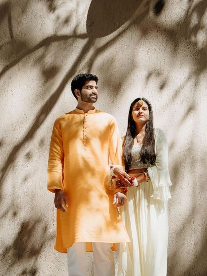 A beautifully lit portrait of a couple in traditional Haldi outfits, with artistic shadows playing on the wall behind them.