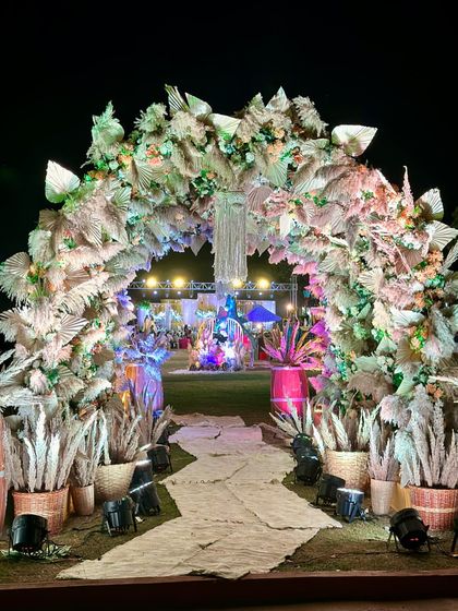 A grand entrance arch designed with a bohemian theme. The structure is covered in dried pampas grass and palm leaves, with a central macrame hanging, creating a dramatic and stylish welcome for a rustic-themed event.