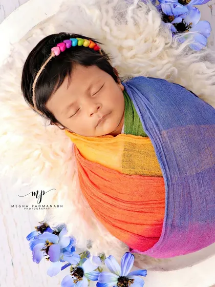 A baby wrapped in a rainbow swaddle, resting on a fluffy white rug with blue flowers.