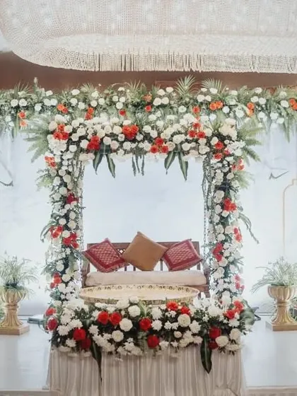 An elegant jhoola (swing) setup for a pre-wedding ceremony, framed by a beautiful arch of red and white flowers. The traditional brass lamp (samai) and golden pots add to the richness of the decor.