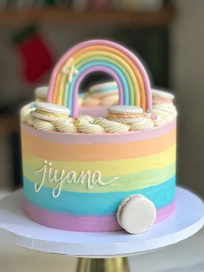 A colorful pastel rainbow cake for a 4th birthday. This design features smooth rainbow buttercream stripes and is topped with a fondant rainbow and delicate macarons.