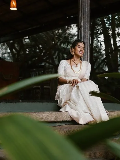 A bride in a white saree sits on the steps of the verandah, a quiet, contemplative moment captured amidst the greenery.