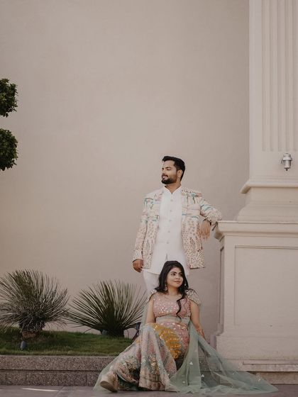 An elegantly styled couple poses against a modern, minimalist building. Their ornate, coordinated outfits bring a touch of royalty to the contemporary setting.