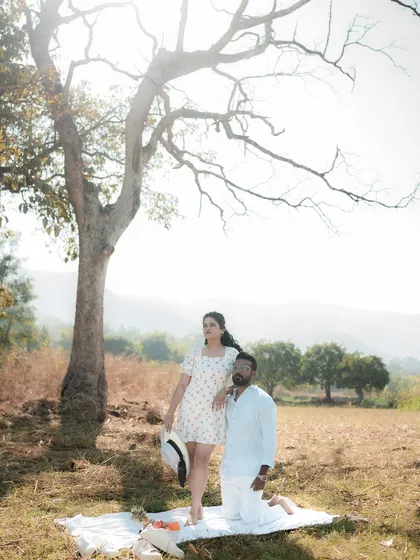 A beautiful wide shot of a couple enjoying a picnic in a sunlit field, framed by a large tree. This image captures the romantic and serene experience of an outdoor couple photoshoot, perfect for a save-the-date announcement.