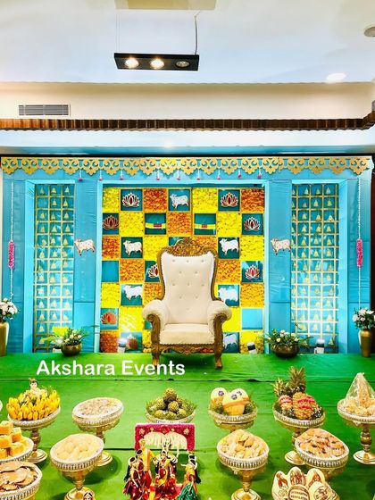 A bright and cheerful Seemantham stage with a blue and yellow theme. The backdrop features a checkerboard pattern with cow and lotus motifs, complemented by a full display of offerings.