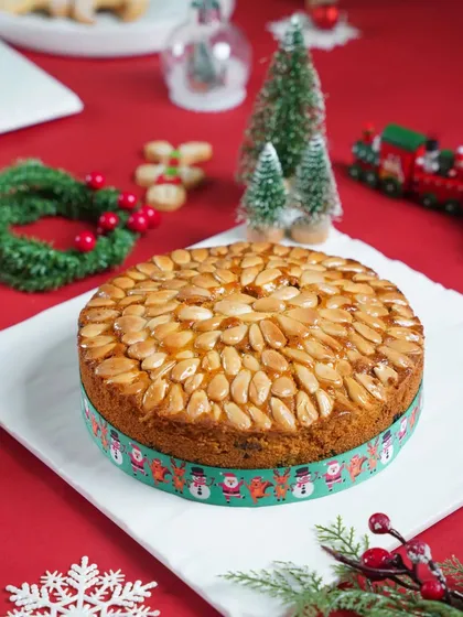 A traditional almond-topped Christmas cake on a white plate, surrounded by festive red and green decorations. The bright, clean lighting gives it a fresh and celebratory look.