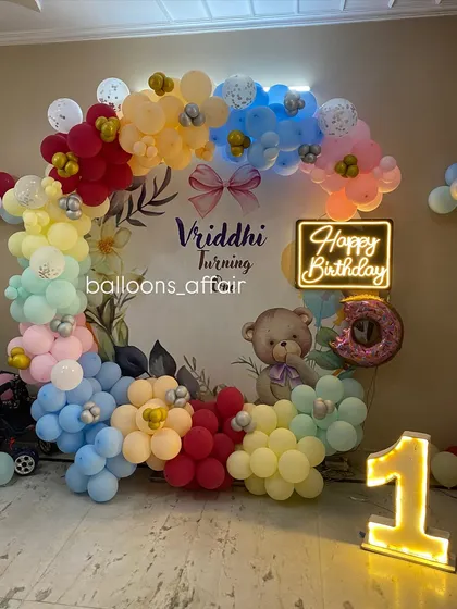 A colorful first birthday decoration with a teddy bear theme. The setup includes a rainbow-colored balloon arch, a floral backdrop, and a light-up number '1'.