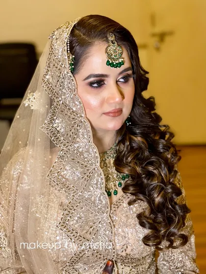 A beautiful portrait of a bride in a beige and gold outfit. The makeup is classic and elegant, with soft curls and a perfectly set dupatta.
