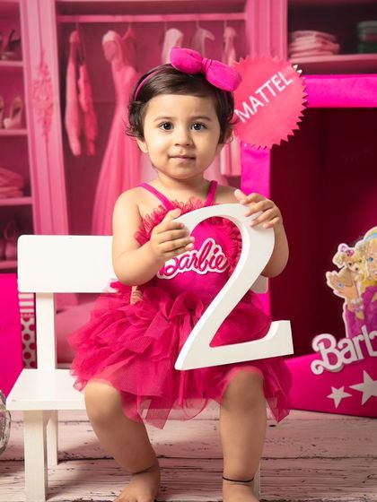 Celebrating turning two with a Barbie theme. This little girl proudly holds up the number '2' in her pink dress, surrounded by disco balls and Barbie accessories.