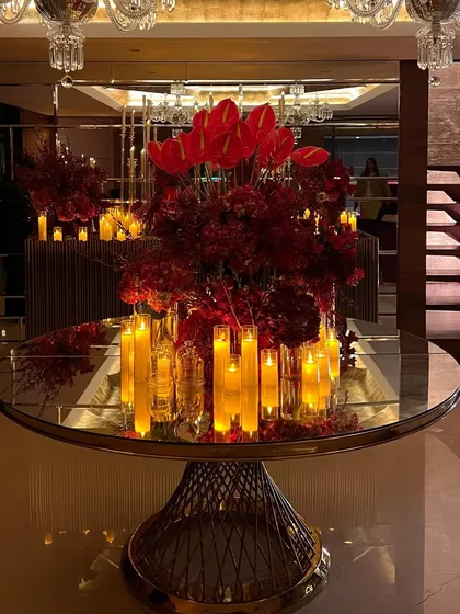 The reflection of the grand red floral centerpiece on the mirrored tabletop. The composition, featuring red anthuriums and glowing candles, is perfectly symmetrical and visually striking.