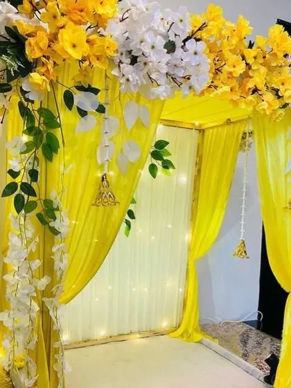 A classic and cheerful four-post canopy mandap, perfect for a traditional Ganpati celebration. The structure is draped in bright yellow fabric and adorned with white and yellow flowers, with elegant bell hangings adding a festive touch.