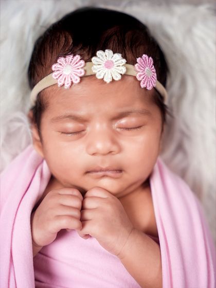A close-up portrait of a newborn's sweet face, adorned with a delicate floral headband. The soft pink wrap complements her beautiful skin tone.