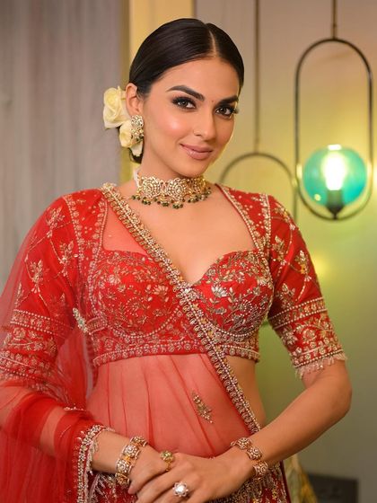 A dazzling bride in a classic red lehenga. The makeup is glamorous yet timeless, with defined eyes and glowing skin, paired with a sleek floral bun.