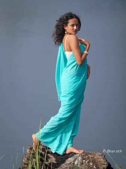 A graceful, full-length shot of the model in the turquoise saree, standing on a log by the water. The pose is elegant and highlights the flow of the fabric.