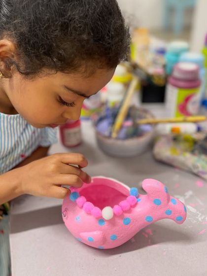 A young girl adds decorative pom-poms to her freshly painted pink whale planter, giving it a touch of personal flair.
