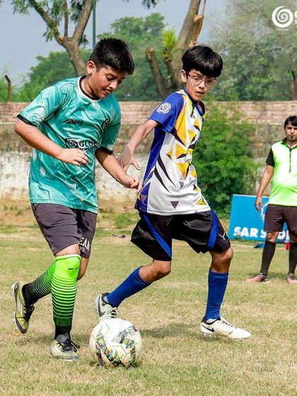 A GSC player shields the ball from an MVTFA opponent, with a coach observing in the background.