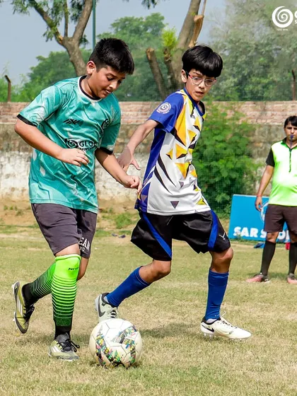 A GSC player shields the ball from an MVTFA opponent, with a coach observing in the background.