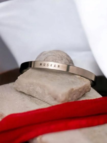 A silver cuff engraved with 'MUSKAN', styled with red and white fabric to create a visually striking product shot.