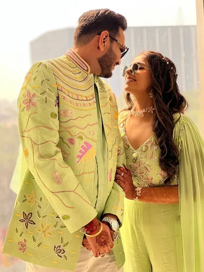 A loving glance between a couple dressed in our coordinated carnival-themed outfits. The intricate, colorful embroidery on his sherwani beautifully matches the floral details on her lime green jumpsuit.