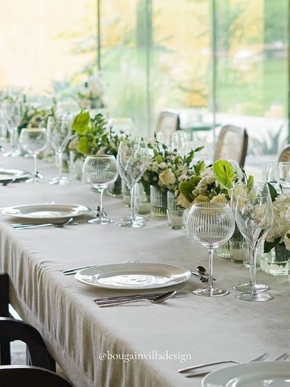 A daytime view of the same long table setup. The white linens and florals look crisp and fresh against the backdrop of the lush green forest seen through the glass walls.