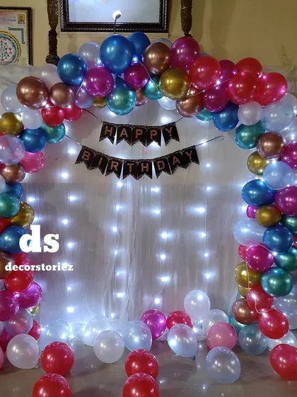 A vibrant and festive balloon arch using multi-colored chrome balloons against a backdrop of white curtains and fairy lights, perfect for any lively celebration.