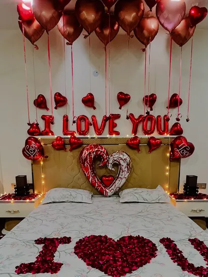 A close-up of the bed decoration, featuring 'I LOVE YOU' foil balloons, chrome and red heart-shaped balloons, and an 'I ❤️ U' message written with fresh rose petals on the bedspread.