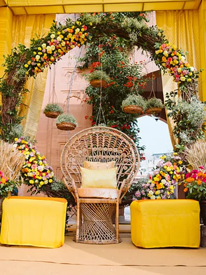A vibrant Haldi setup, featuring a peacock chair framed by a lush arch of yellow and pink florals. The decor is joyful, traditional, and full of color.