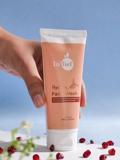 A styled shot of the In'lief face wash with pomegranate seeds scattered in the foreground, hinting at the ingredients. The blue background adds a pop of color and a feeling of freshness.