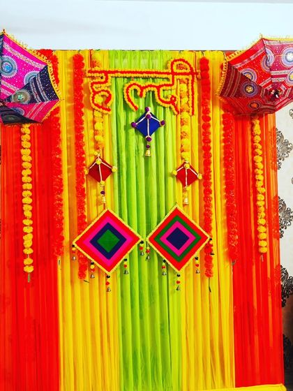 A colourful indoor Haldi backdrop with red, yellow, and green drapes, decorated with marigold garlands and traditional umbrellas.